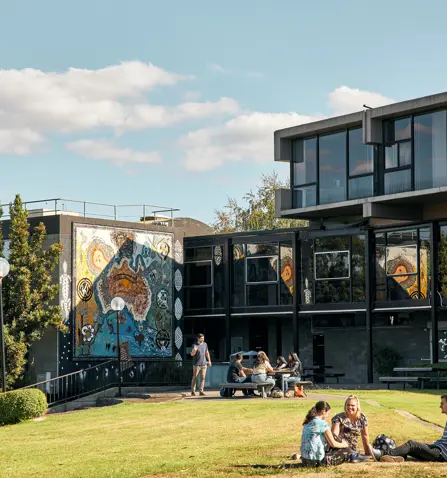 Photo of Gippsland campus from outside. There are people sitting on the grass in the foreground.