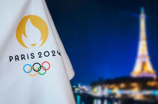 Photo of a Paris 2024 Olympics flag in the foreground with a blurred Eiffel Tower in the background.