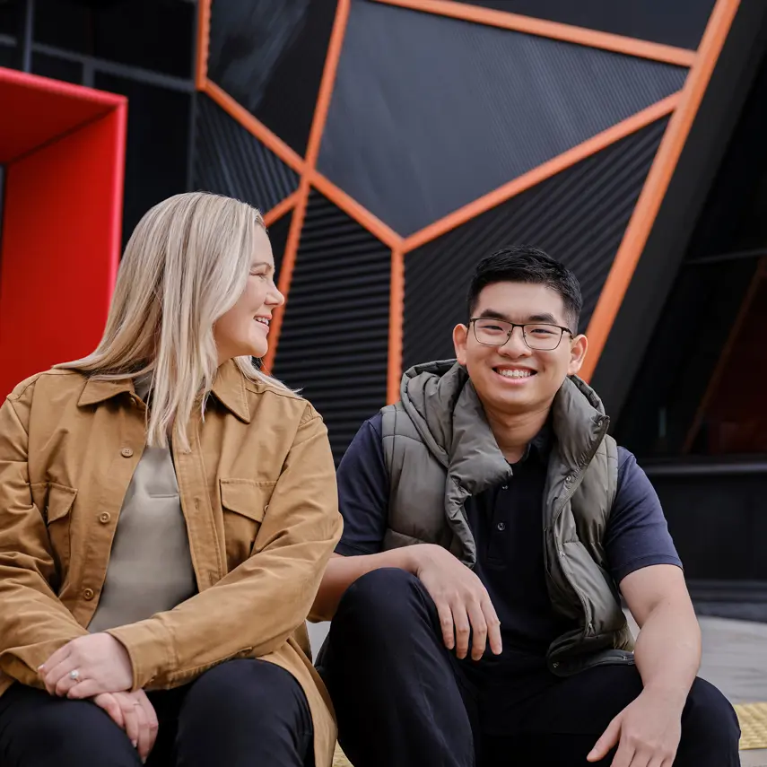 Photo of two smiling people sitting outside in front of a modern building.