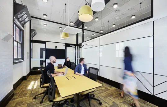 Photo of classroom setting with three people sitting at a big desk and one person near a whiteboard.