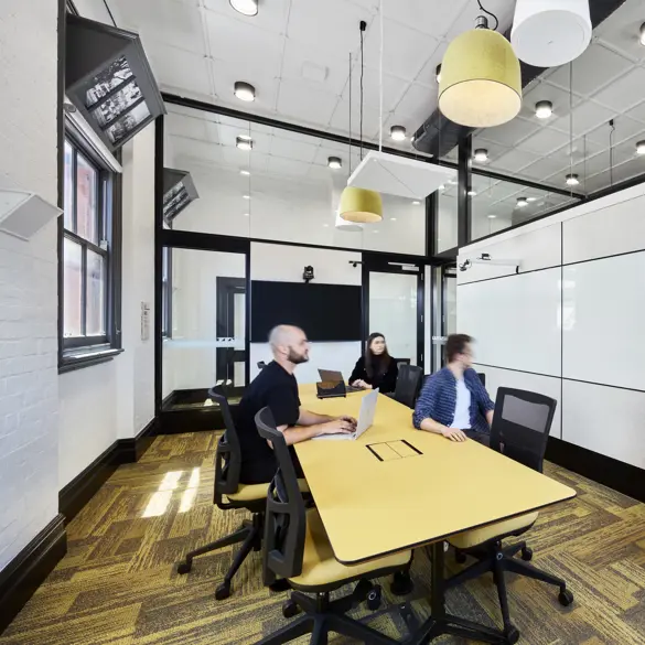 Photo of classroom setting with three people sitting at a big desk and one person near a whiteboard.