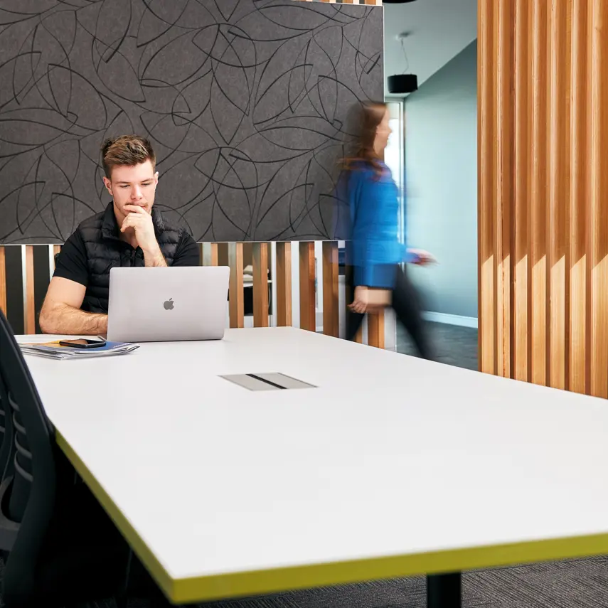Photo of a young man sitting at a desk using a computer. A blurred woman is leaving the room through a door to the right.