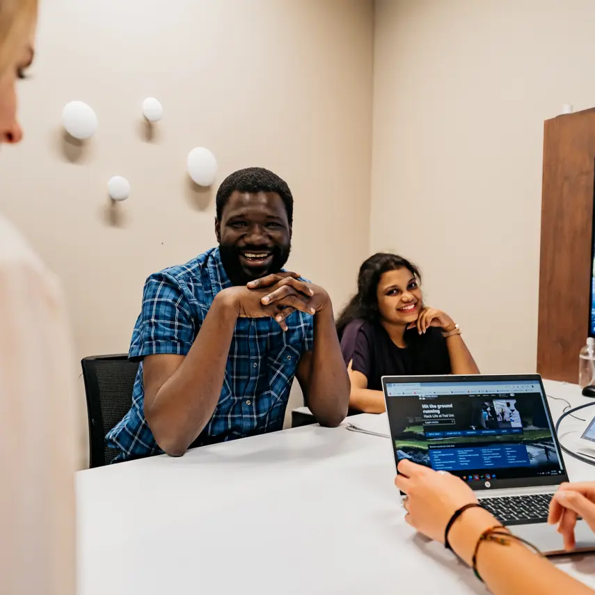 Two students sitting at a table smiling. A laptop and hands can also be seen.